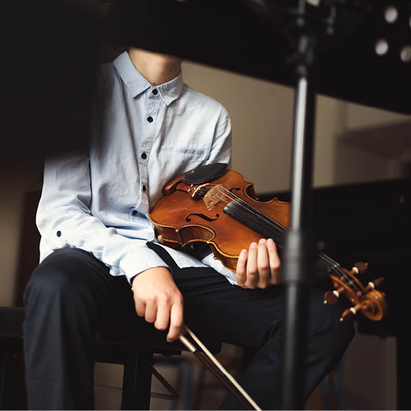 Unrecognizable teenage violinist sitting and holding instrument in his lap, resting between playing. Kid violin player indoors. 