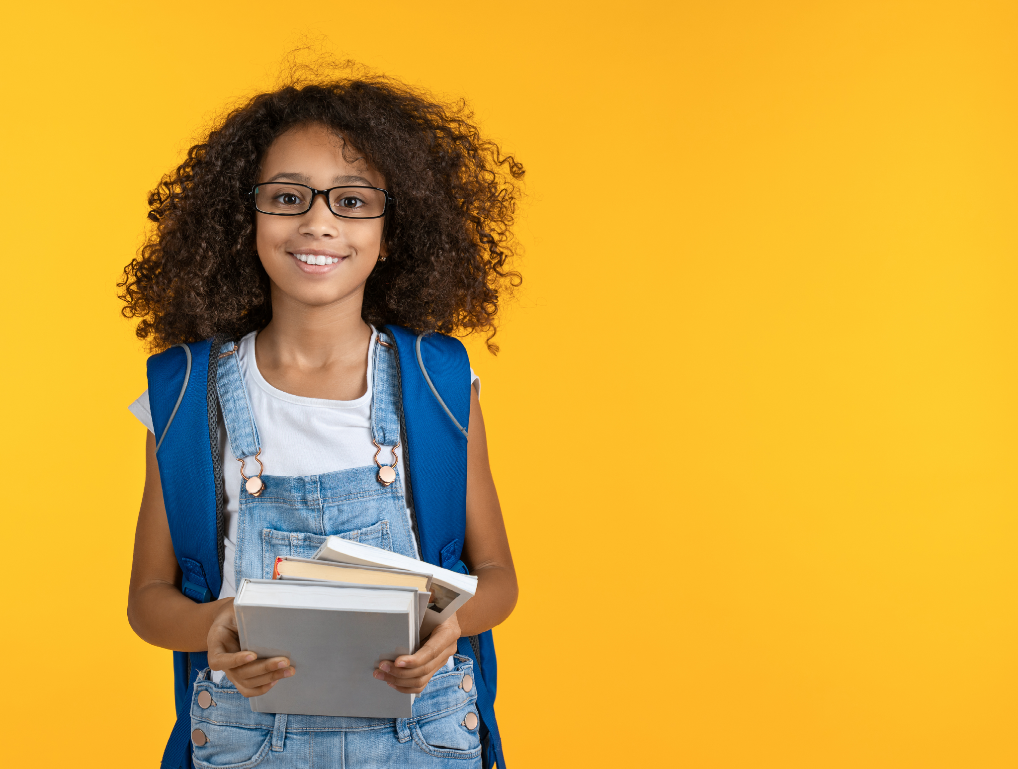 Cheerful young african girl kid in eyeglasses holding notebook and books for study isolated over yellow background