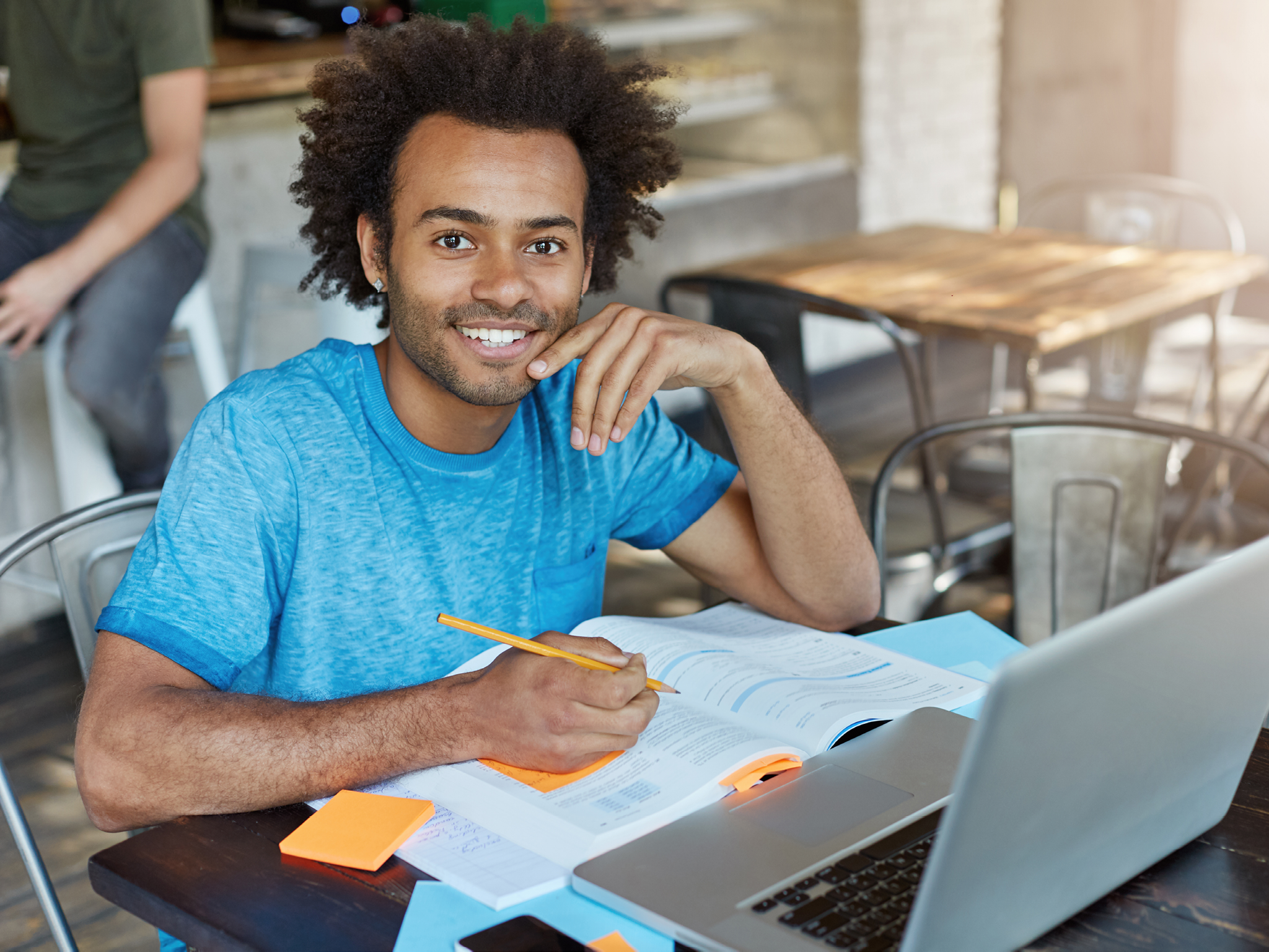 Handsome male student with dark skin, curly bushy hair and bristle smiling broadly at camera demonstrating his perfect white teeth doing his homework while sitting at university canteen using laptop