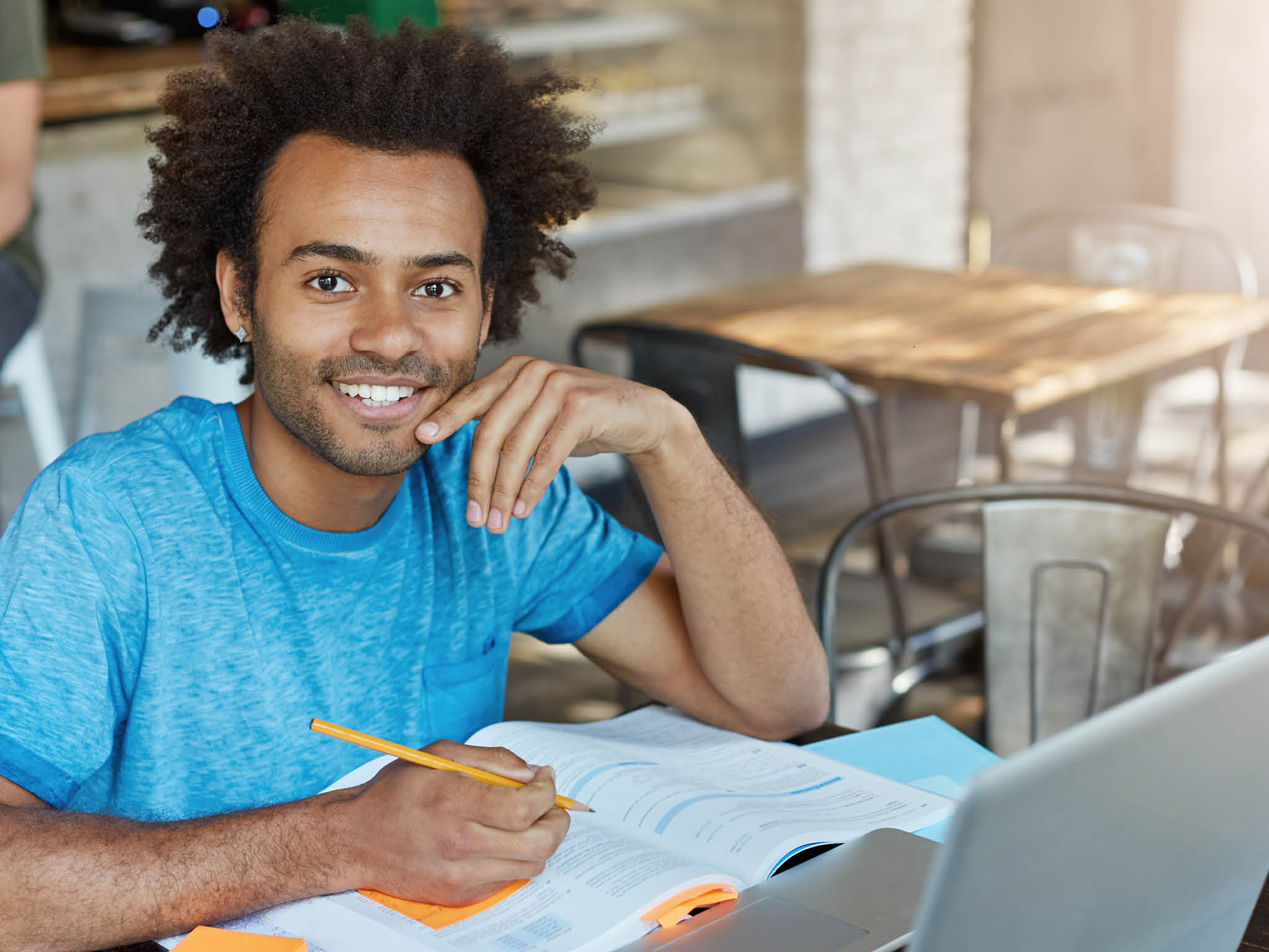 Handsome male student with dark skin, curly bushy hair and bristle smiling broadly at camera demonstrating his perfect white teeth doing his homework while sitting at university canteen using laptop